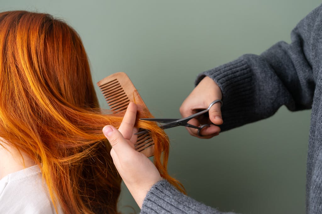 Close-up of red hair being trimmed with scissors and comb on green background.