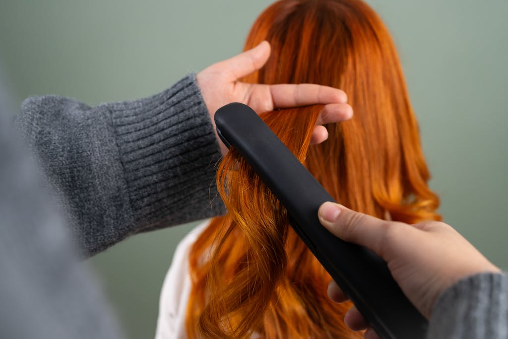 Hands styling long red hair using a flat iron indoors. Haircare and beauty concept.